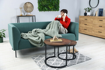 Fototapeta premium Woman with cup of hot drink talking over phone on sofa at home