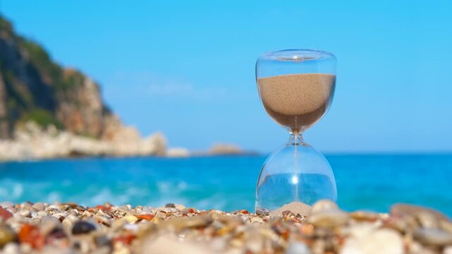 Hourglass measuring time on a sunny beach. Golden sand pouring through a classic hourglass on a pebble beach, with the turquoise sea and blue sky in the background, symbolizing the passage of time