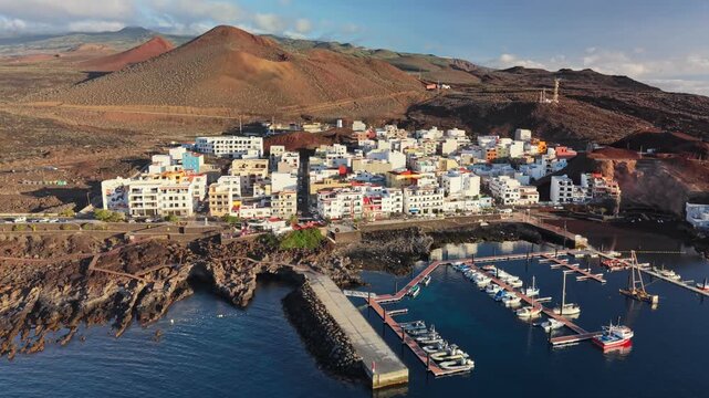 Wide panorama of La Restinga fishing port and coastal town nestled in a volcanic bay. Dramatic drone flyover showing turquoise waters of the Atlantic and black lava fields, El Hierro, Canary Islands