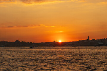 Sunset across the Bosphorus toward Eminonu and Karakoy, with silhouettes of Suleymaniye and Fatih Mosques and the Galata Tower against orange sky in Istanbul, Turkey