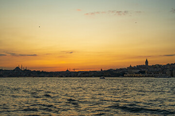 Naklejka premium Sunset across the Bosphorus toward Eminonu and Karakoy, with silhouettes of Suleymaniye and Fatih Mosques and the Galata Tower against orange sky in Istanbul, Turkey