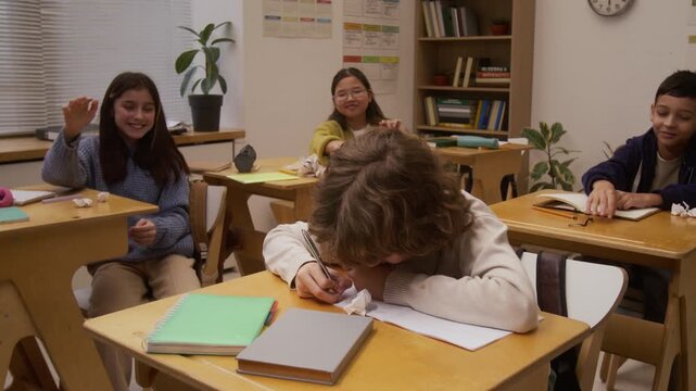 Medium shot of children bullying young boy in school and throwing crumpled paper at him, laughing
