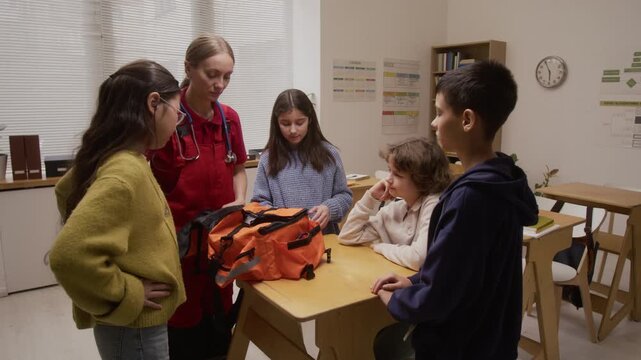 Medium shot of group of children watching interactive presentation with young woman as female paramedic showing contents of medical bag on career day in school