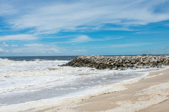 Rompeolas de piedra en playa tropical con espuma de mar blanca bajo cielo azul.