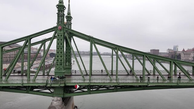 Aerial pulls back from the green steel latticework of the Liberty Bridge (Szabads&aacute;g h&iacute;d) in Budapest, revealing the tram tracks, pedestrians, and the grey waters of the Danube River