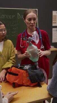 Vertical shot of young woman as female paramedic holding AMBU bag and explaining first aid to children on career day in school