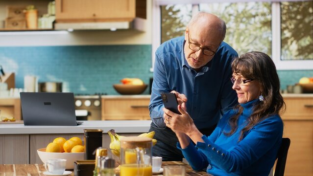 Senior woman and her husband laughing and checking smartphone at the breakfast table to enjoy a cozy meal with toast and coffee. Chatting and reflecting warm retirement lifestyle. Camera B.