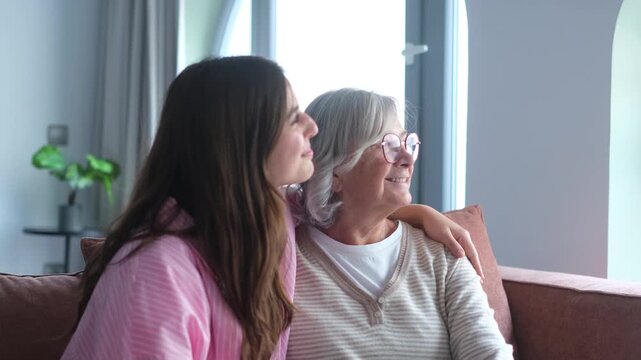 Dreamy pensive young woman hugging older mother smile looking away, feel happy, enjoy priceless and carefree time together at home, recollect, having nostalgic mood, daydreaming seated on sofa indoor