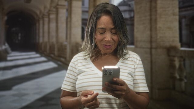 Hispanic middle age woman holding smartphone, thumb tapping screen and smiling while standing in an arched stone building corridor; curiosity connection.