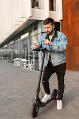 Young and handsome man with electric scooter using smartphone standing against the background of office buildings. Using gadgets in work and everyday life.