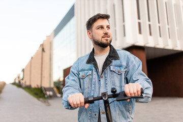 Attractive man in a denim jacket riding electric kick scooter at office buildings background. Driving an ecological mode of transport.