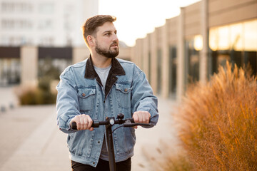 Attractive man in a denim jacket riding electric kick scooter at office buildings background. Driving an ecological mode of transport.