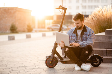 Young and handsome man with a beard sitting on electric scooter on the street and works at his laptop at sunset. Freelance work outside the home. Driving an ecological mode of transport.