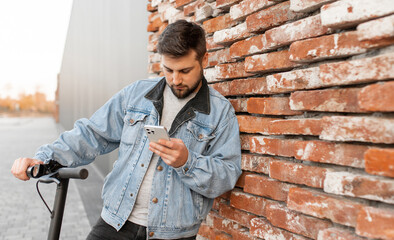 Man wearing a denim jacket on electric scooter uses smartphone against the background of brick wall. Person uses smartphone to search for geolocation. 
