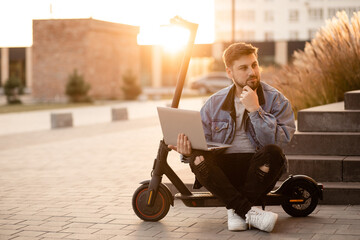 Young and handsome man with a beard sitting on electric scooter on the street and works at his laptop at sunset. Freelance work outside the home. Driving an ecological mode of transport.