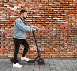 Attractive man in a denim jacket riding electric kick scooter at  brick wall background. Driving an ecological mode of transport.