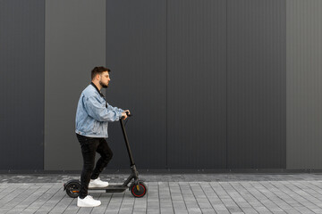 Attractive man in a denim jacket riding electric kick scooter on a grey background of a street building. Driving an ecological mode of transport.