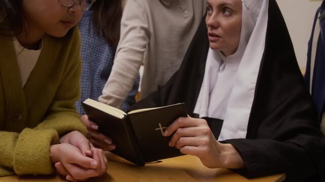 Close up shot of young woman as Catholic nun holding bible and reading religious texts to group of children during interactive presentation on career day in school classroom