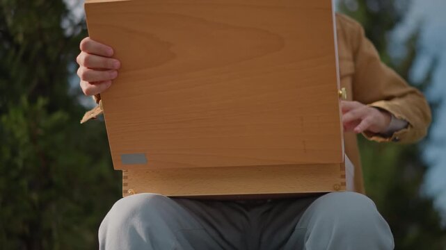 portable easel case opens on lap, hands releasing latches and lifting wooden lid, sunlight filtering through trees, focused preparatory moment for outdoor painting session, compact gear reveal