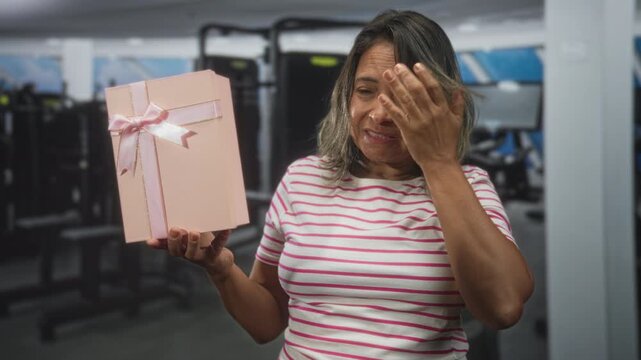 Woman holds pink gift box with ribbon and covers face with hand inside a building fitness gym with visible machines; surprise reflection.