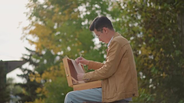 young artist sketching on bench with wooden easel box open, casual jacket, focused hands drawing in sketchbook, autumn trees and soft sunlight, peaceful streetside park atmosphere showing creative