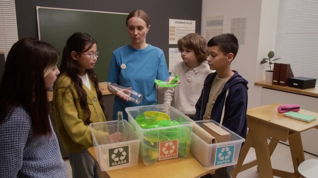 Medium shot of young woman as environmental educator teaching waste sorting with group of children watching during interactive presentation on career day in school
