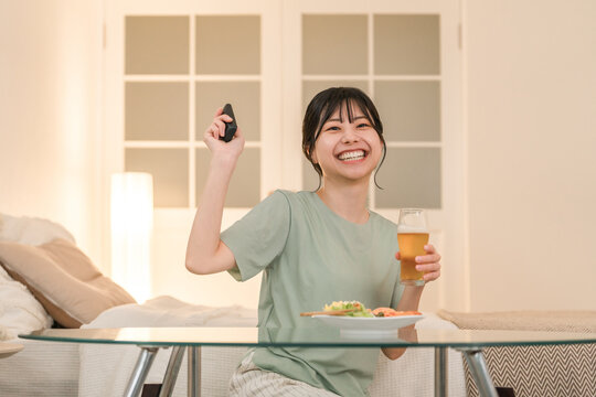 Young Asian woman laughing while watching TV and having a drink at dinner