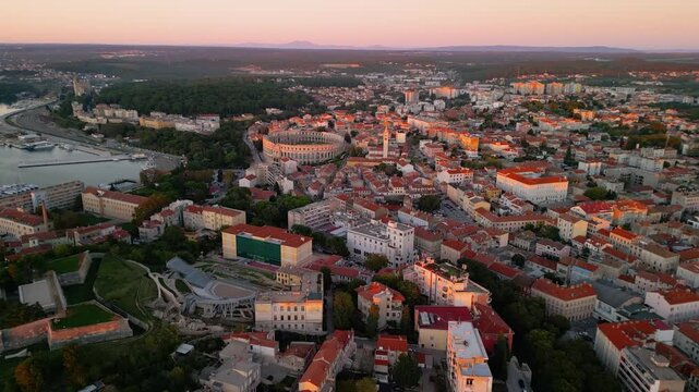 Epic Pula arena sunset aerial reveal. 
Cinematic 4K drone pull-away flight 
from historic Roman amphitheater 
and harbor at twilight in Croatia.
