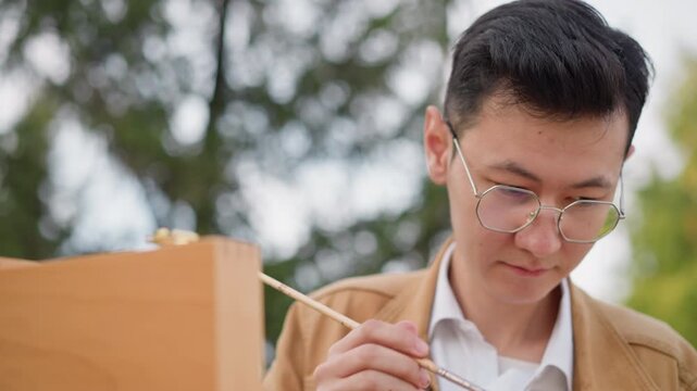 Asian man painting on easel outdoors, focused portrait study in leafy park, wearing glasses and brown jacket, sketching on canvas, adjusting brush, examining reference, closeup framing captures