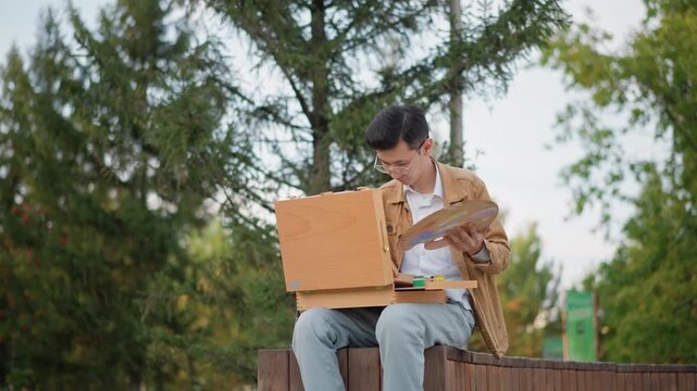 park easel illustrator studying color palette young creator reviewing reference and mixing paints on wooden palette, seated on bench under tall trees, careful brush strokes and compositional study,