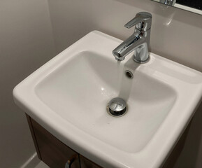 Fototapeta premium A stream of water flows from a chrome single-lever faucet into a small, white ceramic sink mounted on a wooden vanity cabinet.