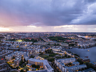 Obraz premium Urban beauty and tranquility of Stockholm with illuminated buildings, parks, and waterways under a dramatic cloudy sky at dusk, seen from an aerial perspective
