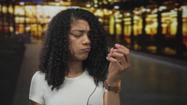 Woman holding broken headphone cable on a dimly lit city street at night with a disgusted frown; frustration.