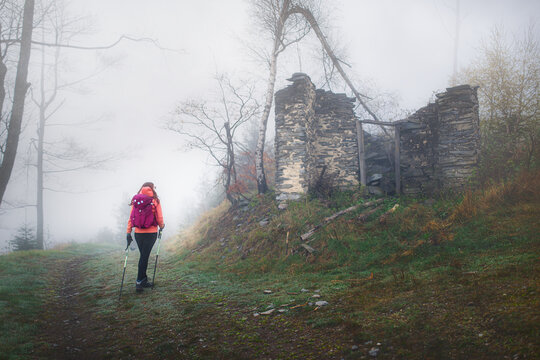 Female hiker with backpack and trekking poles walking on a trail past abandoned stone ruins in a thick foggy forest.