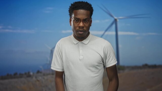Man wearing white polo shirt and glasses points finger to camera in front of windmill building; confidence.