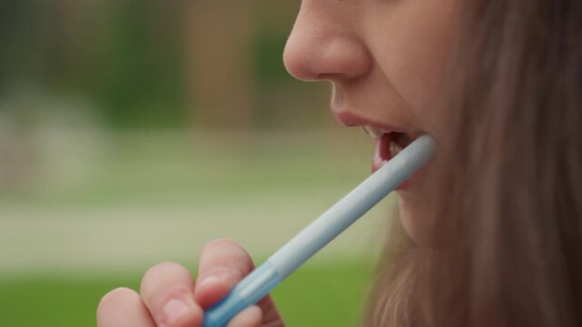 young female student chewing pencil outdoors, close profile showing lips and hand holding pen, green blurred campus background, contemplative nervous habit while studying, soft natural light
