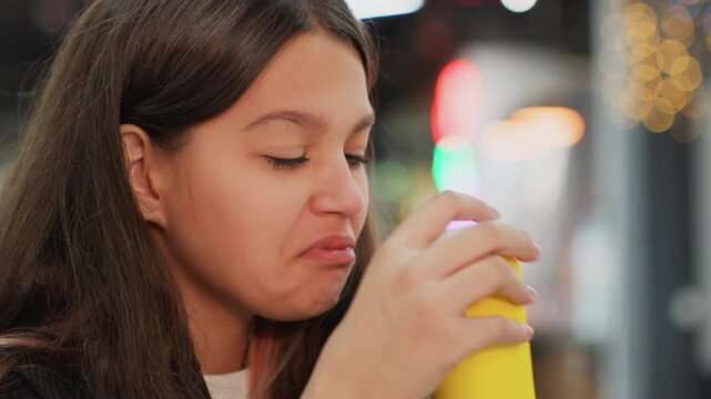 student sipping drink in cafeteria, candid closeup capturing first sip reaction. female student tastes beverage from yellow cup, small spill on lip, initial grimace, then soft smile and contented