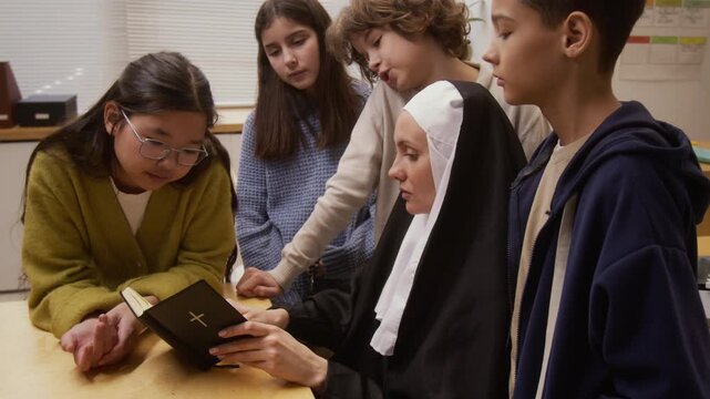 Side view shot of young woman as Catholic nun reading bible to diverse group of children with boy asking question during interactive lecture on career day in school classroom, copy space