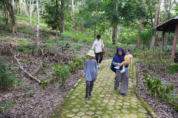 Asian Muslim mother walking with her kids on a path through a lush green park in the morning sun