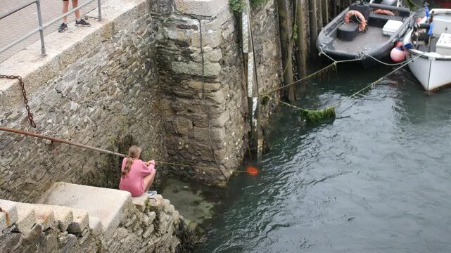 A girl sitting on stone steps beside a harbor wall lowers a crabbing line into the water with moored boats and seaweed covered ropes in Looe Cornwall England