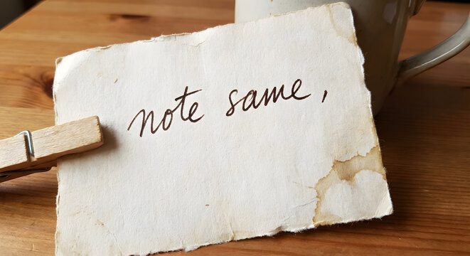 A rustic wooden clothespin gently holds a piece of aged, rough-edged paper with the handwritten phrase "note same," resting on a warm wooden table next to a blurry mug.