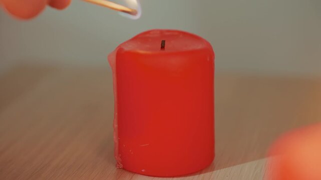 hand striking match to candle, closeup sequence of match ignition and flame catching red pillar wick on wooden table, careful deliberate motion suggests home ritual, aromatherapy setup or prelude