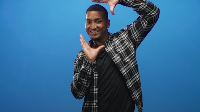 Young black man framing with hands against a blue studio backdrop, smiling at camera; playful confidence.
