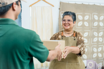 Obraz premium Elderly asian artisan online seller gives parcel box to courier, completes small business shipping handoff, home studio shibori fabric tie-dye brand delivery pickup, friendly customer service.