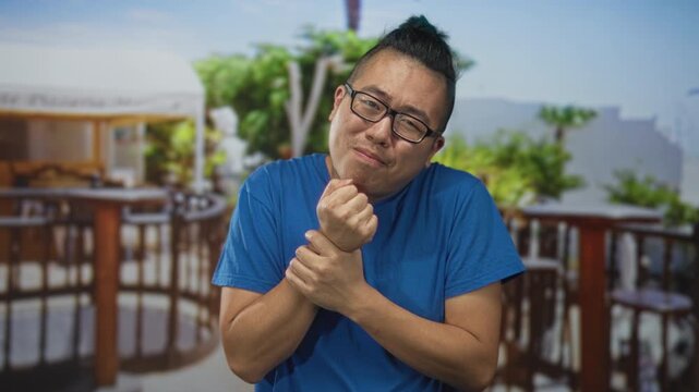 Young chinese man wearing glasses and a blue t shirt clutches his wrist with both hands on a restaurant terrace near a building, squinting and pressing fingers; discomfort.