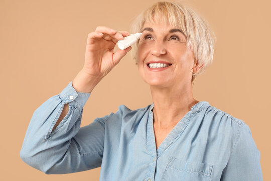 Mature woman using eye drops on beige background