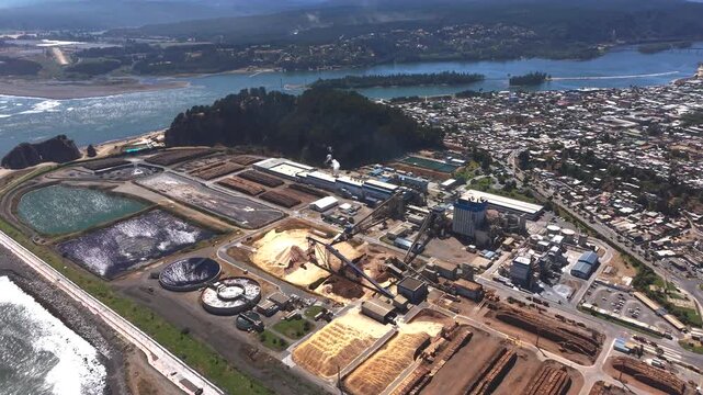 Wide aerial angle of a massive industrial pulp mill complex located on the coastline near the river mouth in Constituci&oacute;n, Chile, showing wood stockpiles and factory layout.