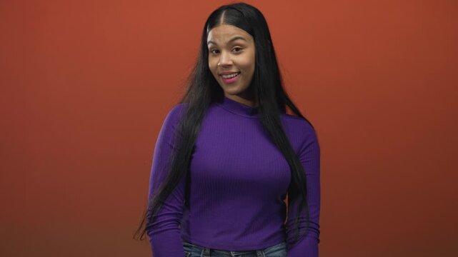 Standing young latin woman making ok sign with fingers in orange studio wearing purple top and jeans; approval consent affirmation agreement.