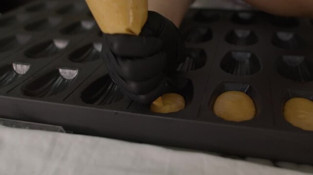 close-up shot of a pastry chef's hand in a black glove using a piping bag to fill a dark silicone mold with smooth cake batter on a marble surface