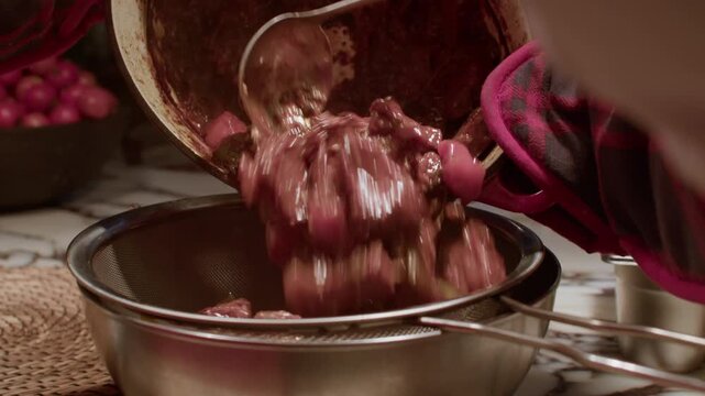 A detailed close-up shot of a person pouring a rich, dark red stew with large meat chunks and vegetables from a pot into a metal strainer